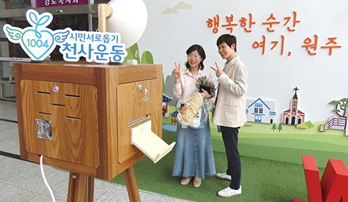 고객 두 명이 이날 프리마켓과 함께 열린 ‘1004 시민서로돕기 천사운동’ 이벤트에 참여해 폴라로이드 사진을 찍으며 웃고 있다. 방금 산 뻥튀기와 싱싱한 생화를 손에 들고 V자를 그려 보인다.